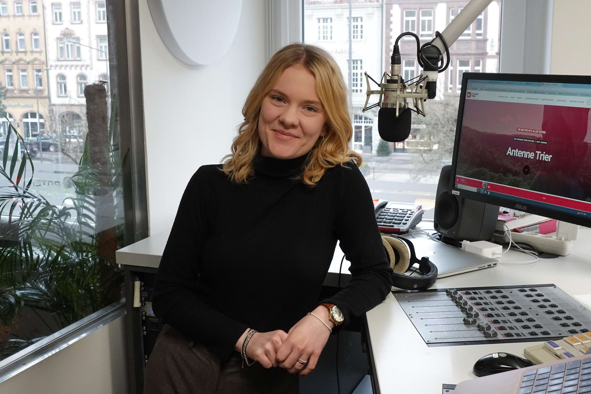 Junge Frau mit blonden, lockigen Haaren sitzt an einem Tisch in einem Büro. Im Hintergrund ein Monitor und Mikrofon.