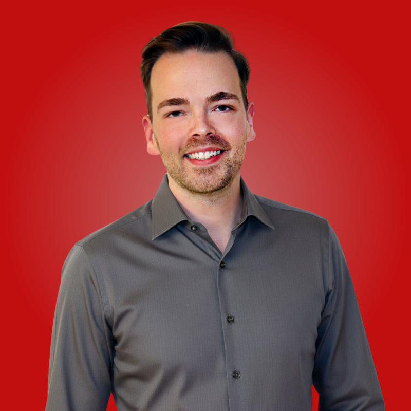 Ein junger Mann mit lächelndem Gesicht trägt ein graues Hemd vor einem roten Hintergrund.